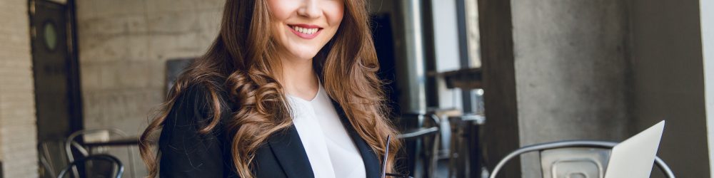 Widely smiling businesswoman working on laptop sitting in a cafe. Woman with long hair wears black jacket and white blouse and holds black eyeglasses in hands.